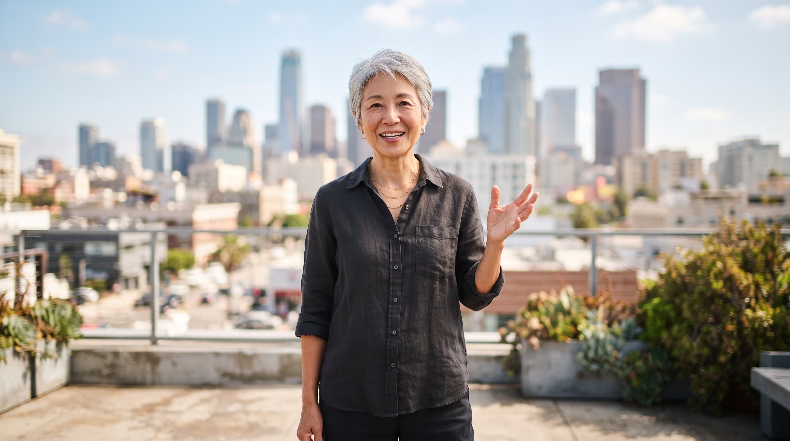 Silver-haired woman on a sunny city rooftop gesturing as she talks to the camera